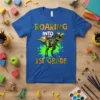 Roaring Into 1st Grade T-Shirt with a green T-Rex dinosaur wearing goggles and a jetpack, printed on a blue shirt