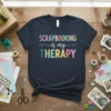 Scrapbooking is my Therapy quote in colorful, playful typography on a navy t-shirt, surrounded by craft supplies