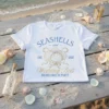 White t-shirt with blue and gold text "Seashells and Wedding Bels Bachelorette Party Est. 2025" and seashell graphic