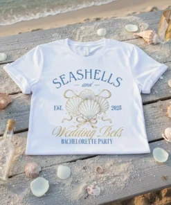 White t-shirt with blue and gold text "Seashells and Wedding Bels Bachelorette Party Est. 2025" and seashell graphic