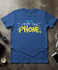 Blue t-shirt with yellow and white text reading "shhh, i'm on The PHONE." in a playful font