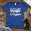 Blue t-shirt with white text: "what i ask for: snuggles, what i get: struggles" repeated, featuring small graphic icons