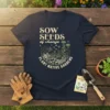 Sow Seeds of Change Plant Native Gardens text with floral and butterfly graphics on a navy t-shirt