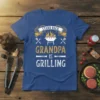 Stand Back Grandpa Is Grilling text design with barbecue grill graphic and utensils, on a blue t-shirt
