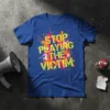 Royal blue t-shirt with yellow, orange, and pink paint splatter graphic and bold yellow text "STOP PLAYING THE VICTIM"