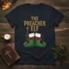 The Preacher Elf Christmas T-Shirt featuring golden text and green elf boots with red bows