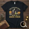 This Witch Needs Coffee Before Any Hocus Pocus text with pumpkins, black cat, and cauldron graphic on a black t-shirt
