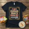 Black t-shirt with "Tiny Human Snack Dealer #nannylife" text in colorful gradient and white font, framed by ornate gold border