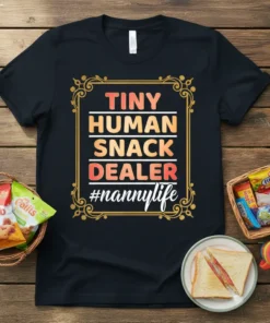 Black t-shirt with "Tiny Human Snack Dealer #nannylife" text in colorful gradient and white font, framed by ornate gold border
