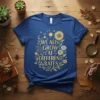 Blue t-shirt with golden text "We All Grow At Different Rates" surrounded by sunflowers and floral elements