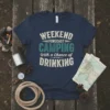 Weekend Forecast Camping With A Chance Of Drinking text design on a navy t-shirt, featuring vintage typography