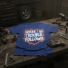 Blue t-shirt with white text that reads "WHERE I GO TROUBLE FOLLOWS" in a bold, jagged font, outlined in orange