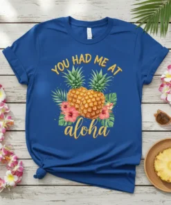 Blue t-shirt with yellow text "YOU HAD ME AT aloha" and two pineapples with hibiscus flowers and palm leaves